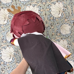 May include: A plush doll with burgundy hair, a gold crown, and a black cape. The doll has a white collar and pink accents. The background features a floral wallpaper design.