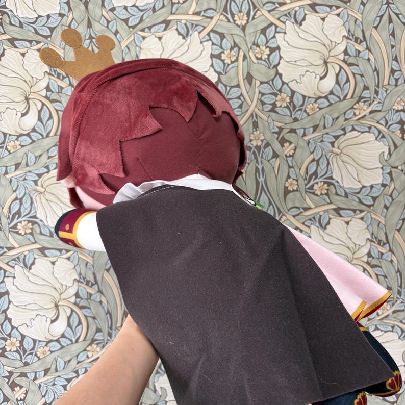 May include: A plush doll with burgundy hair, a gold crown, and a black cape. The doll has a white collar and pink accents. The background features a floral wallpaper design.