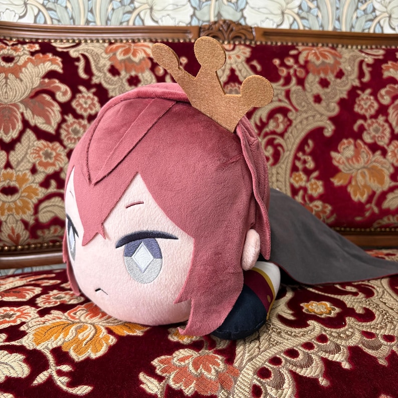 May include: A plush doll with a stylized anime-inspired design. The doll features a pink head with a brown crown, a gray cape, and a detailed face with a diamond-shaped eye. The doll is lying on a red floral patterned surface.