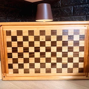 May include: A rectangular wooden cutting board with a checkerboard pattern. The board is made of light and dark wood squares, framed by a lighter wood border. The board is approximately 18 inches long and 12 inches wide.