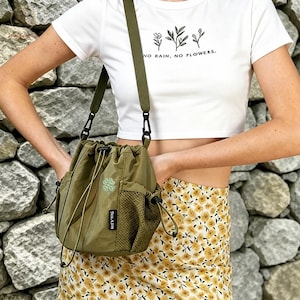 Könnte beinhalten: Olivgrüne Crossbody-Tasche mit Kordelzug, Netztasche und kleiner Stickerei. Die Tasche hat einen langen, verstellbaren Riemen. Das weiße Crop-Top hat den Text "NO RAIN, NO FLOWERS."