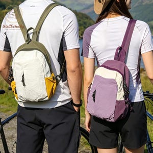 May include: Two small backpacks are shown, one olive green and cream, the other purple and cream. They are worn by people near bicycles. The backpacks have a minimalist design with a single main compartment and a small front pocket.