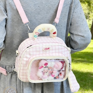 Puede incluir: Una mochila rosa y blanca a cuadros con correas rosas y un asa blanca. La mochila tiene un bolsillo frontal transparente que muestra muñecos de peluche. El asa tiene bordada la palabra "BABY".
