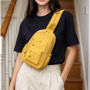 May include: Mustard yellow sling bag with multiple zipped compartments and an adjustable strap. The bag features a small front pocket and a top handle. The bag is worn across the body.