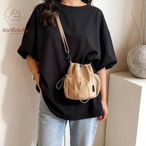 May include: A beige drawstring bucket bag with a shoulder strap. The bag features two front pockets and a mesh side pocket. The bag is worn over a black t-shirt and blue jeans.