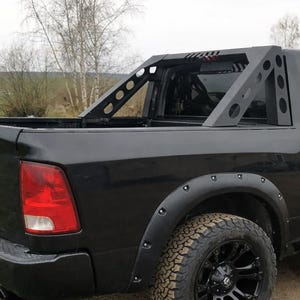 May include: Black pickup truck with a custom roll bar. The roll bar is made of black metal with a geometric design. The truck has black wheels and tyres, and black fender flares. The background shows a field and trees.