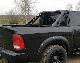 Op de afbeelding: Zwarte pick-up truck met een aangepaste rolbeugel. De rolbeugel is gemaakt van zwart metaal met een geometrisch ontwerp. De truck heeft zwarte wielen en banden en zwarte spatbordverbreders. De achtergrond toont een veld en bomen.