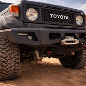 Peut inclure: Un véhicule tout-terrain Toyota noir avec de gros pneus robustes est garé sur une surface sablonneuse. L'avant du véhicule présente une calandre noire avec le logo Toyota, des phares ronds et un pare-chocs robuste avec un treuil.