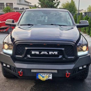 May include: A dark blue RAM pickup truck with a black grille and bumper. The word "RAM" is prominently displayed on the grille. The truck has bright headlights and a license plate. Red tow hooks are visible on the bumper.