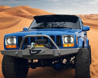 May include: A blue Jeep Cherokee SUV with a gray front bumper and a winch, parked on sand dunes. The vehicle has large, black off-road tires and the word "Jeep" is visible on the front.
