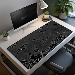 May include: A black and white desk mat with a topographic design. The mat is rectangular and covers a large portion of the desk. The design features white lines on a black background, creating a visual texture. A computer monitor, lamp, and plants are also on the desk.