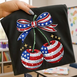 Könnte beinhalten: Schwarzes T-Shirt mit einem patriotischen Kirsch- und Schleifendesign. Die Kirschen und die Schleife sind mit roten, weißen und blauen Strasssteinen und goldenen Sternen verziert. Das Design wird von einer Hand gehalten.
