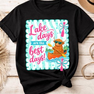 Könnte beinhalten: Schwarzes T-Shirt mit einer farbenfrohen Grafik. Die Grafik zeigt eine Cartoon-Hochlandkuh mit Sonnenbrille, die ein Kanu paddelt. Der Text lautet "Lake days are the best days!" in Pink und Blau.