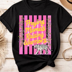 Könnte beinhalten: Schwarzes T-Shirt mit dem Text "Praying Mamas Raising Warriors" in gelber Schrift. Das Design umfasst einen rosa gestreiften Hintergrund, florale Akzente und eine weiße Tigerillustration.