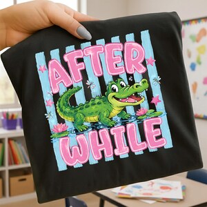 Könnte beinhalten: Schwarzes T-Shirt mit einem Cartoon-Alligator-Design. Die Worte "AFTER WHILE" sind in Rosa gehalten, vor einem blau gestreiften Hintergrund. Der Alligator ist grün mit gelbem Bauch und befindet sich im Wasser mit rosa Lotusblumen.