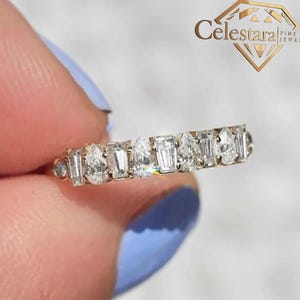 Può includere: Un anello d'oro con una serie di diamanti a taglio pera e smeraldo alternati. L'anello è tenuto da una persona con lo smalto blu. L'anello è di Celestarajewelry.