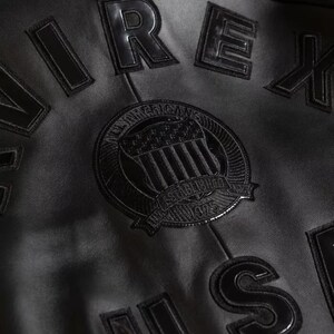 Puede incluir: Primer plano de una chaqueta de cuero negra con letras y parches en relieve. La chaqueta presenta la palabra "AIREX" en letras grandes en relieve, junto con un parche circular que dice "All American" y "Established 1975."