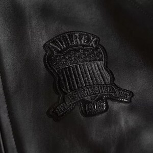 Puede incluir: Primer plano de una chaqueta de cuero negra con un emblema texturizado. El emblema, en forma de escudo, tiene la palabra "AVIREX" en la parte superior y "ESTABLISHED 1975" en la inferior. El cuero de la chaqueta tiene una sutil veta.