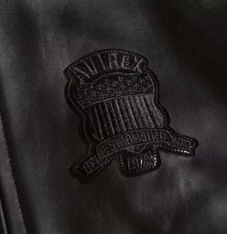 Puede incluir: Primer plano de una chaqueta de cuero negra con un emblema texturizado. El emblema, en forma de escudo, tiene la palabra "AVIREX" en la parte superior y "ESTABLISHED 1975" en la inferior. El cuero de la chaqueta tiene una sutil veta.