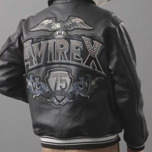Könnte beinhalten: Schwarze Lederjacke mit weiß-grauem Streifenbund. Auf der Rückseite befindet sich ein Adleremblem über dem Wort "AVIREX" und ein "75"-Wappen. Die Jacke hat einen klassischen Bomberstil.