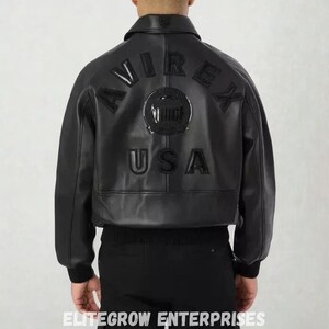 Puede incluir: Chaqueta bomber de cuero negro con la palabra "AVIREX" arqueada en la espalda, encima de un emblema circular y la palabra "USA". La chaqueta tiene dobladillo y pu&ntilde;os de canal&eacute;. Un estilo cl&aacute;sico.