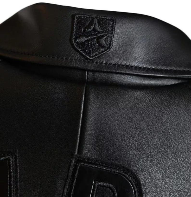 Puede incluir: Primer plano de una chaqueta de cuero negra. La chaqueta presenta un peque&ntilde;o emblema bordado en el cuello y letras grandes cosidas en la parte inferior. El cuero tiene un acabado liso y brillante. La chaqueta es una prenda de vestir exterior elegante.