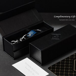 May include: A black gift box with a silver keychain inside. The keychain has a blue leather strap and a silver metal charm. The box has the text "Complimentary Gift Box" and "Included with every item" printed on it.