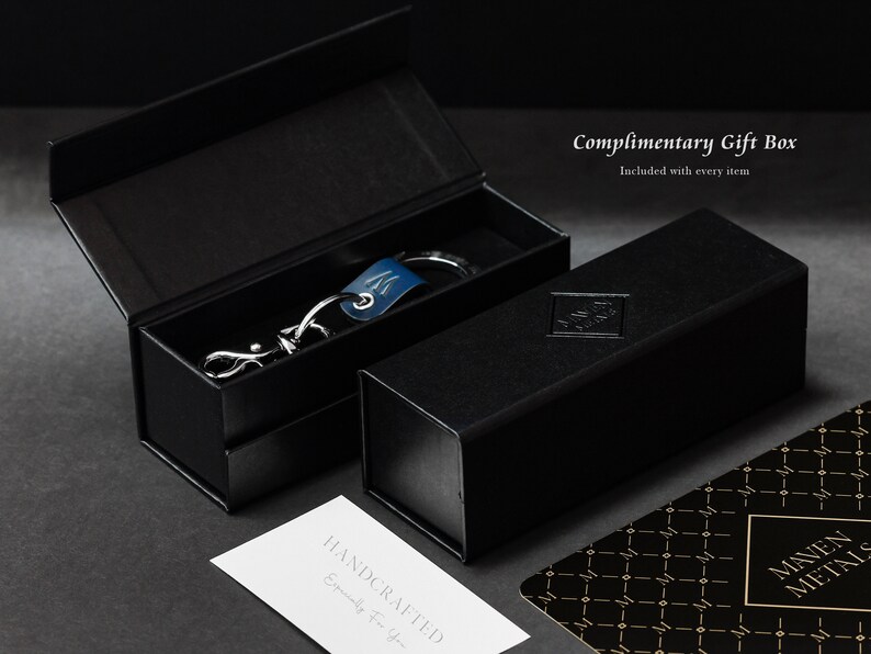 May include: A black gift box with a silver keychain inside. The keychain has a blue leather strap and a silver metal charm. The box has the text "Complimentary Gift Box" and "Included with every item" printed on it.