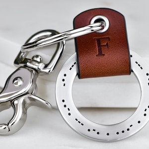May include: A silver key chain with a brown leather strap. The strap has the letter "F" embossed on it. The keychain has a silver ring with a series of dots around the outside.