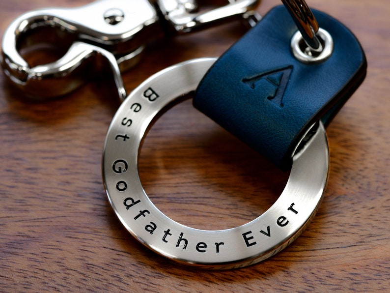 May include: A silver keychain ring engraved with "Best Godfather Ever" text. The ring is attached to a navy blue leather strap with the letter "A" embossed on it. A silver clasp is also attached.