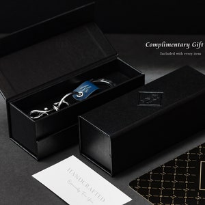 Op de afbeelding: Een zwarte cadeaudoos met een zilveren sleutelhanger erin. De sleutelhanger heeft een blauwe leren band en een zilveren metalen hanger. De doos is gelabeld met 'Complimentary Gift Box' en 'Included with every item'.