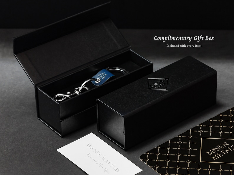 Op de afbeelding: Een zwarte cadeaudoos met een zilveren sleutelhanger erin. De sleutelhanger heeft een blauwe leren band en een zilveren metalen hanger. De doos is gelabeld met 'Complimentary Gift Box' en 'Included with every item'.