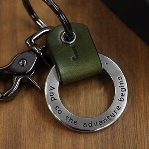 Op de afbeelding: Een groene leren sleutelhanger met een zilveren ring en een zilveren sluiting. De ring heeft de tekst "And so the adventure begins" gegraveerd.