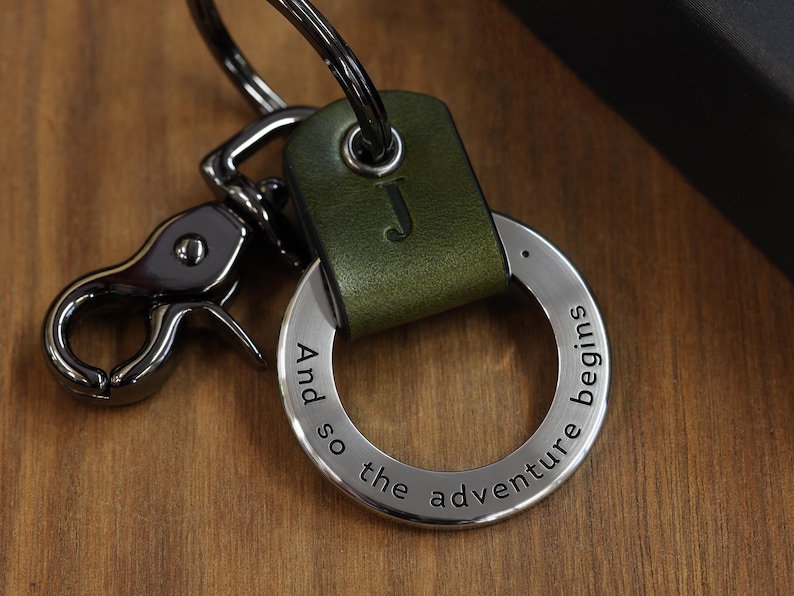 Op de afbeelding: Een groene leren sleutelhanger met een zilveren ring en een zilveren sluiting. De ring heeft de tekst "And so the adventure begins" gegraveerd.
