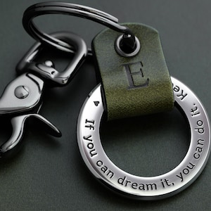 Op de afbeelding: Een zilveren sleutelhanger met een groene leren band en de letter "E" erop gegraveerd. De sleutelhanger heeft een ronde ring met de inscriptie "Als je het kunt dromen, kun je het doen."