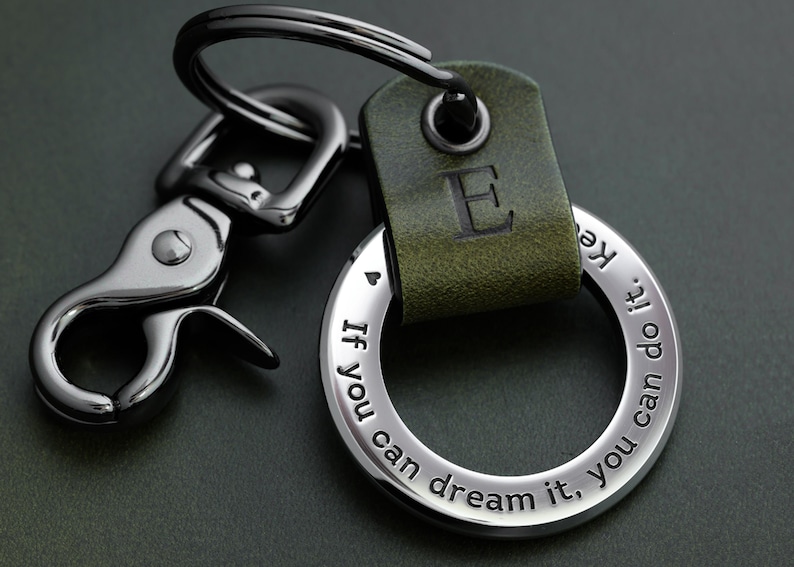Op de afbeelding: Een zilveren sleutelhanger met een groene leren band en de letter "E" erop gegraveerd. De sleutelhanger heeft een ronde ring met de inscriptie "Als je het kunt dromen, kun je het doen."