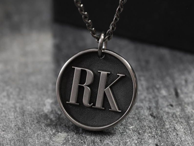 May include: A silver chain necklace with a round pendant featuring the initials "RK" in raised lettering on a black background.