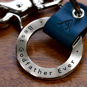 Op de afbeelding: Een zilveren sleutelhanger met de woorden "Best Godfather Ever" gegraveerd. De ring is bevestigd aan een blauwe leren sleutelhanger met een gegraveerde zilveren letter "A".