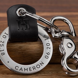 May include: A silver keychain with a black leather strap. The keychain has a round metal tag engraved with the text "CAMERON 06.20.19 1.25.18" and the initial "H".