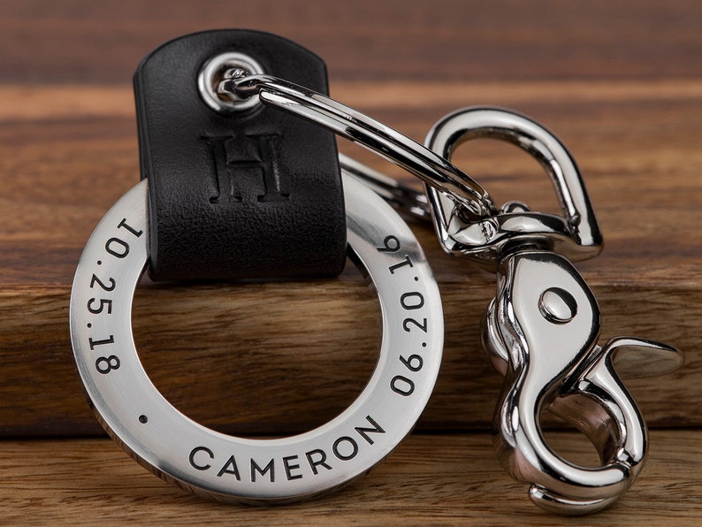 May include: A silver keychain with a black leather strap. The keychain has a round metal tag engraved with the text "CAMERON 06.20.19 1.25.18" and the initial "H".