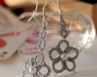 Può includere: Un paio di orecchini fatti a mano di colore argento. Ogni orecchino presenta un design a forma di fiore con una perla al centro. Gli orecchini sono sospesi da ganci per orecchie argentati. Lo sfondo è sfocato.