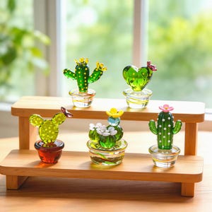 May include: A collection of six glass cactus figurines in various shapes and colors, each in a small glass pot. The cacti are green with yellow, pink, and white flower accents. They sit on a two-tiered wooden display shelf.