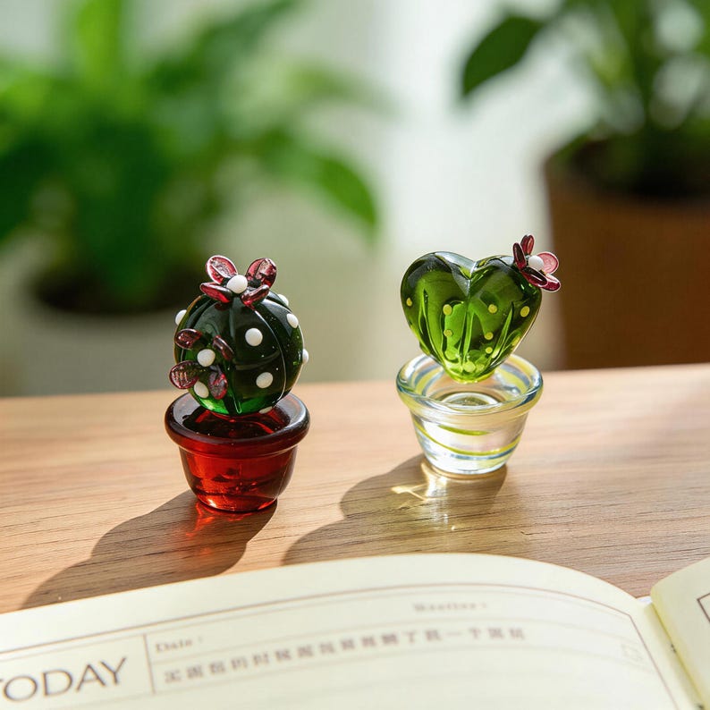 May include: Two glass cactus figurines. One is round with white dots and red flowers, set in a red pot. The other is heart-shaped with yellow dots and a pink flower, in a clear pot. Both are on a wooden surface.