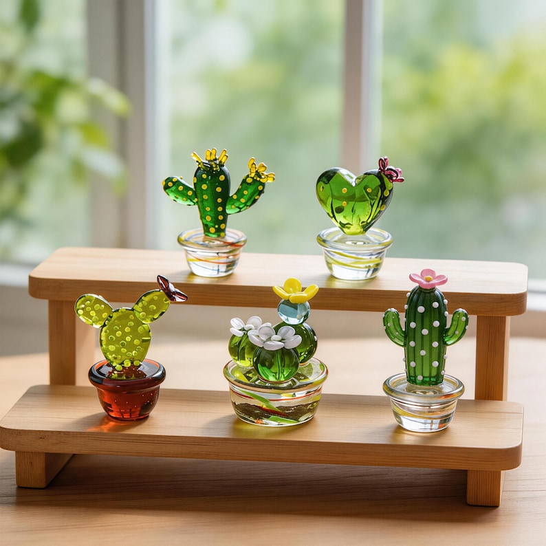 May include: A collection of six glass cactus figurines in various shapes and colors, displayed on a two-tiered wooden shelf. The cacti are green with yellow, pink, and white flower accents, and sit in small glass pots.