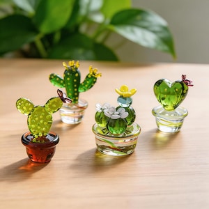 May include: A collection of miniature glass cactus sculptures in various shapes and colors. The cacti are green with yellow and white accents, and are displayed in small, decorative pots. The background is a blurred view of green foliage.