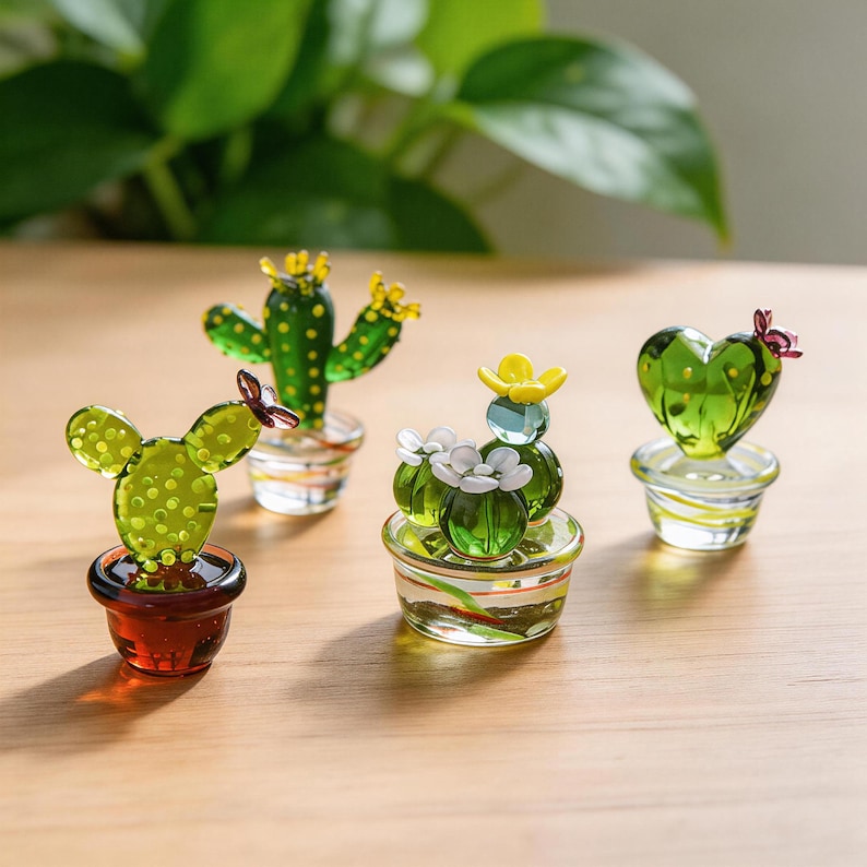 May include: A collection of miniature glass cactus sculptures in various shapes and colors. The cacti are green with yellow and white accents, and are displayed in small, decorative pots. The background is a blurred view of green foliage.