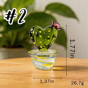 May include: A glass cactus sculpture in a heart shape, with yellow dots and a pink flower. The sculpture is in a clear pot with blue and yellow stripes. It measures 1.77 inches tall and 1.37 inches wide, and weighs 26.7 grams. The text "#2" is in the upper left corner.