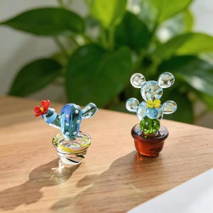 May include: Two glass cactus figurines. One is blue with white polka dots and a red flower, the other is a bear-shaped cactus with a yellow flower. Both are in small pots, set on a wooden surface.