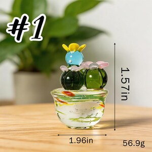 May include: A glass cactus sculpture with three cacti on top of a clear bowl with fish inside. The sculpture is 1.57 inches tall and 1.96 inches wide. The sculpture weighs 56.9 grams. The text "#1" is in the upper left corner.