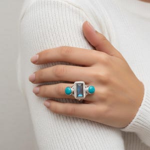 Könnte beinhalten: Ein silberner Ring mit einem rechteckigen blauen Edelstein und zwei türkisfarbenen Steinen. Der Ring hat eine seilartige Textur um den zentralen Stein. Der Ring wird an einer Hand getragen.